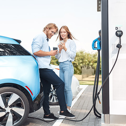Transportation. Young couple traveling by car having stop at charging station boyfriend drinking coffee talkingn with girlfriend browsing smartphone joyful while waiting vehicle to charge Young couple man and woman traveling together by new car having stop at charging station boyfriend holding cup drinking hot coffee talking with girlfriend browsing smartphone smiling joyful while waiting vehicle to charge fully